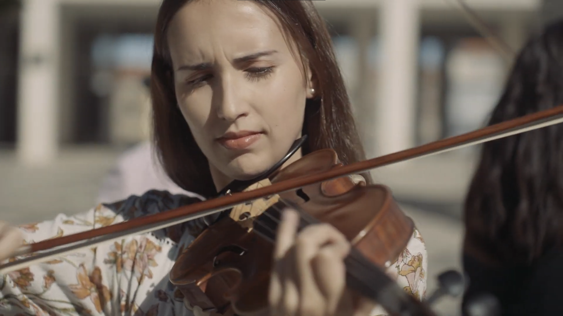 Plano apertado de uma rapariga a tocar violino. Ela tem cabelo liso e veste uma roupa florida. A imagem tem tons térreos.